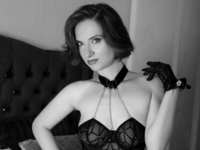 Black-and-white photo of EllyMuller with short dark hair, wearing a sheer black bustier with chains, collar, and black gloves, showing her tits and a confident, sexy expression.