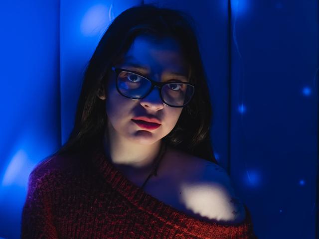 Isabella Rossi, avec ses cheveux foncés, ses lunettes, son pull rouge et une épaule dénudée, regarde l'objectif sous une lumière bleue. Ses seins sont légèrement visibles sous le pull. Elle porte du rouge à lèvres et a une expression sérieuse..