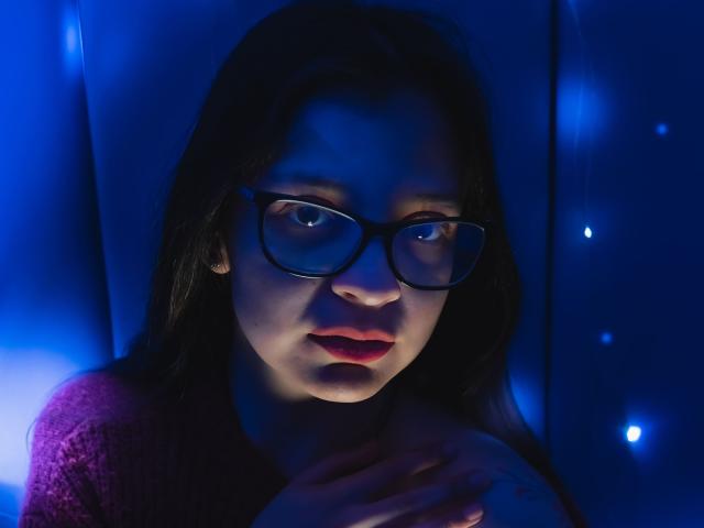 Isabella Rossi, avec ses cheveux foncés, ses lunettes et son rouge à lèvres rouge, tient sa poitrine, a l'air sérieuse, est éclairée par une lumière bleue et porte un pull en tricot violet. Ses seins sont légèrement visibles..