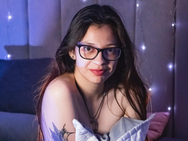 IsabellaRossis with glasses, long brown hair, and a tattoo on her left arm, holding a pillow, looking at the camera with a slight smile, wearing a necklace.