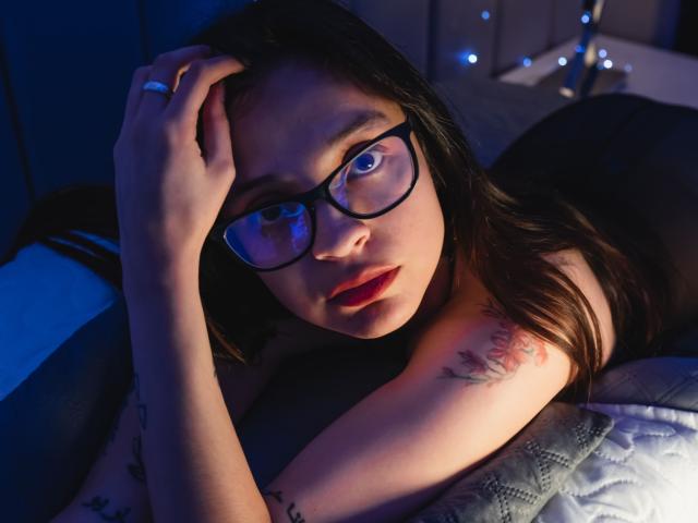 Isabella Rossi, avec des lunettes, allongée sur un lit, une main sur la tête, lumière bleue, tatouage sur le bras, rouge à lèvres rouge, cheveux noirs, regardant la caméra, bague au doigt, lumière douce, couette de lit..