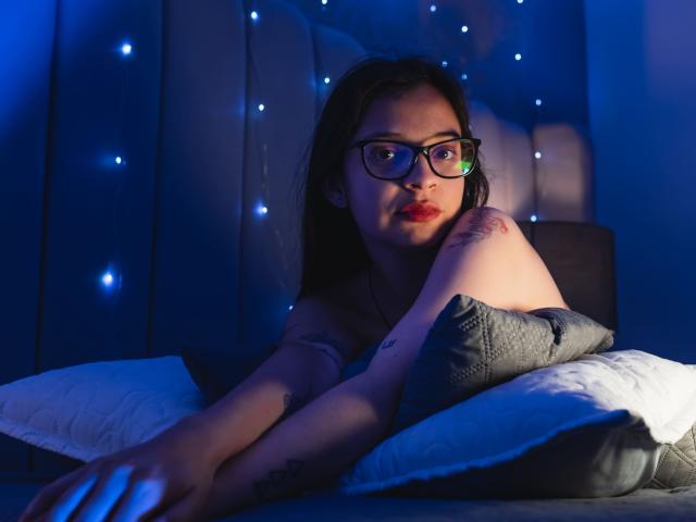 Isabella Rossi, avec des lunettes, du rouge à lèvres et des tatouages, est allongée sur un lit, les bras croisés, tenant un oreiller gris, avec des lumières bleues en arrière-plan, et a l'air légèrement agacée..