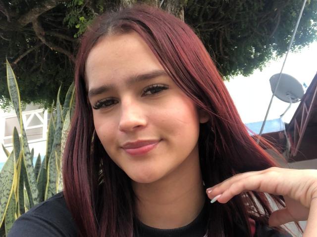 MelodyMansson has red hair, brown eyes, and a slight smile. She's wearing a black shirt, with her hand near her head. Her nails are manicured with white tips.
