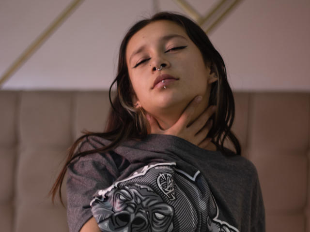 LilithSwett has dark hair, closed eyes, and a nose ring, wearing a gray shirt with a graphic. Her hand grips her neck. She's on a beige couch.