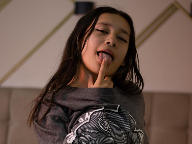 LilithSwett with long brown hair, nose ring, and septum piercing, tongue out, finger in mouth, wearing a grey graphic tee, looking slightly up, with a relaxed expression.