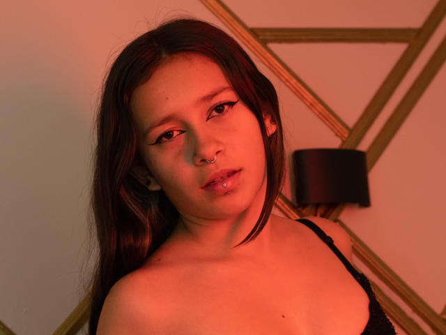 LilithSwett has long brown hair, a nose ring, and a lip piercing, wearing a black strap top, with a red-orange light on her. Her tits are visible.