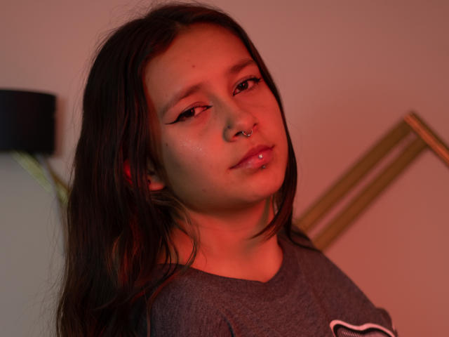 LilithSwett has long dark hair, a nose ring, and winged eyeliner. She's wearing a gray shirt, with red lighting on her face. Her lips are slightly parted.