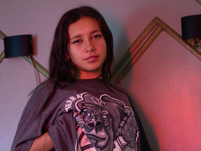 LilithSwett with long brown hair, nose ring, and glossy lips, wearing a gray "Berserker" shirt, looking at camera, soft red and blue lighting.