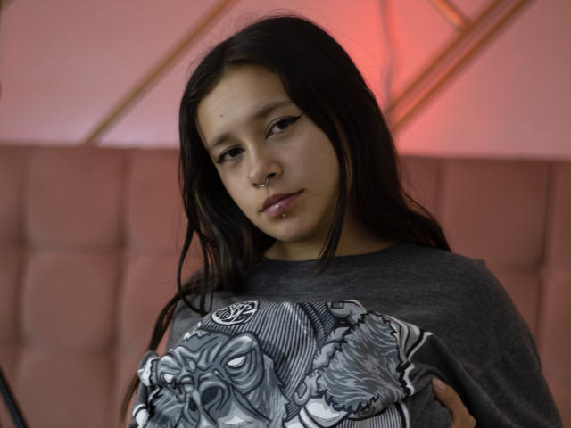 LilithSwett with long black hair, nose ring, and grey graphic tee, holding detailed black-and-white design, looking slightly down with a neutral expression. Soft pink background.