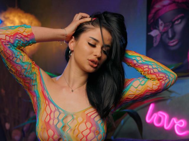 ChanelCharmx, con i capelli scuri, indossa una maglia trasparente, colorata e a maniche lunghe, ha le mani tra i capelli, mostrando il seno e una piccola collana, in una stanza poco illuminata..