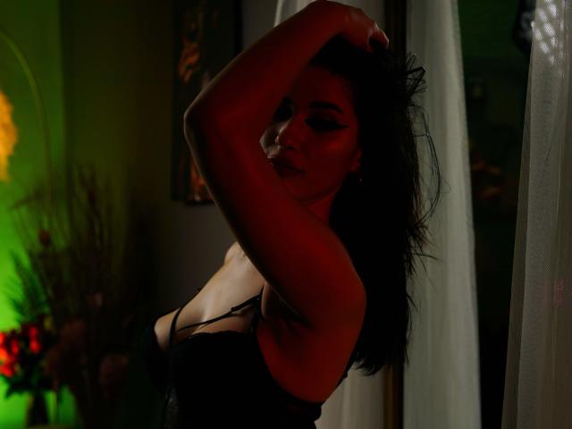 ChanelCharmx with dark hair, raising arm, wearing black lingerie, shadowed face, red light, white curtain, looking seductive, hand in hair, medium shot, dim lighting.