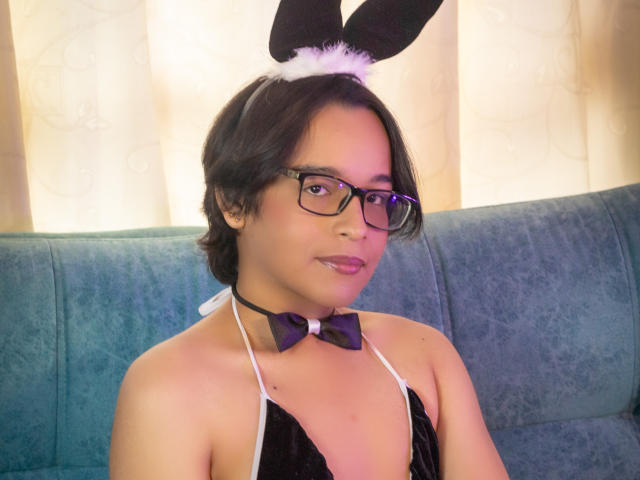 DannyHotz, wearing black and white bunny ears with white fur, glasses, purple bowtie, and black halter top, showing small tits, with short black hair and a cute smile.