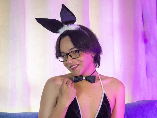 DannyHotz wearing black bunny ears, glasses, and a black halter top with white trim, showing a cute smile, holding a collar with a black bow.