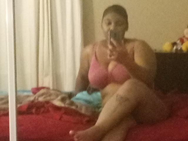 Arissy with big tits in a pink bra, sitting on a red bed, holding a phone, taking a mirror selfie, legs crossed, in a slightly blurry photo.