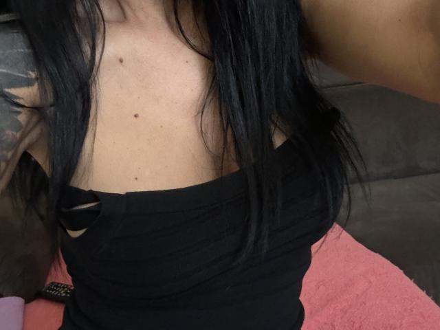 AndraD's black tank top shows her bare shoulders with small moles, long straight black hair, and a tattoo on her left arm. Her small tits are slightly visible.