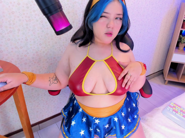 AntonelaGray met blauw en zwart haar, knippend, draagt een rode en gele bikini-top, een blauwe rok met sterrenpatroon en gele armbanden. Haar borsten zijn zichtbaar, haar achterkant is bedekt..