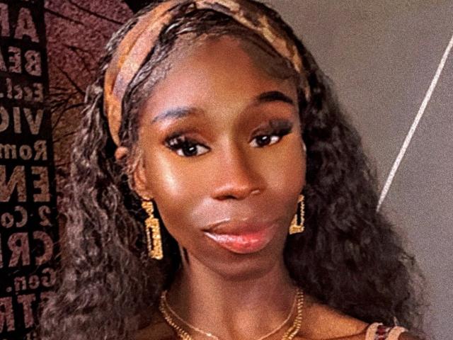 TsHottieX has dark brown skin, curly black hair, and wears a brown headband, gold earrings, and a gold necklace. Her glossy pink lips are slightly parted.
