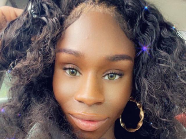 TsHottieX has dark curly hair, green eyes, and wears large gold hoop earrings. She's looking slightly to the side with a subtle smile and smooth brown skin.