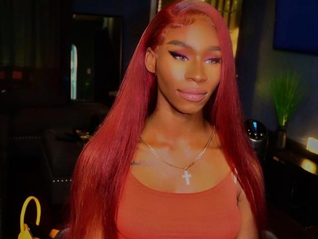 TsHottieX with long red hair, orange tank top, gold necklace with cross, pink eyeliner, and a slight smile. Her brown skin and full lips stand out.