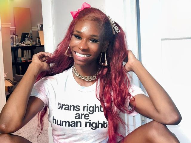 TsHottieX has long red hair, pink bow, diamond hairpiece, big smile, white tee, chain necklace, and earrings, sitting with hands in hair, showing her shiny teeth and cute ass.