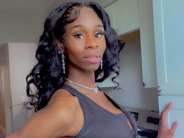 TsHottieX a la peau foncée, de longs cheveux noirs ondulés, et porte un débardeur noir avec un collier de diamants et des boucles d'oreilles pendantes. Elle sourit, avec une légère teinte rose sur les lèvres..