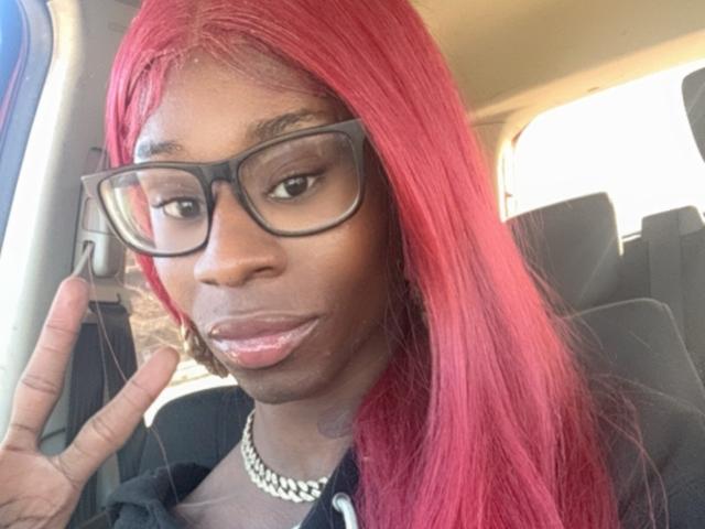 TsHottieX with red hair, black glasses, and a pearl necklace, holding up a finger in a car, showing off glossy pink lips. Boobs not visible. Ass not visible.