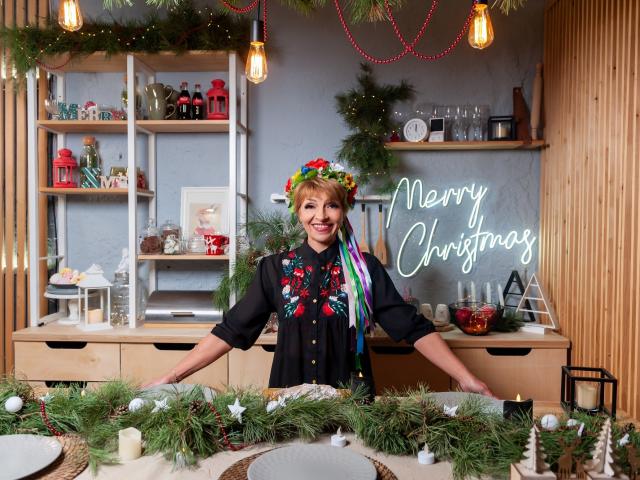 EllaBonito com uma coroa colorida na cabeça, camisa preta com estampa floral vermelha, sorrindo, braços apoiados em um balcão de madeira, decoração de Natal, luzes penduradas, guirlanda verde, velas, prateleiras de madeira com itens vermelhos e brancos..