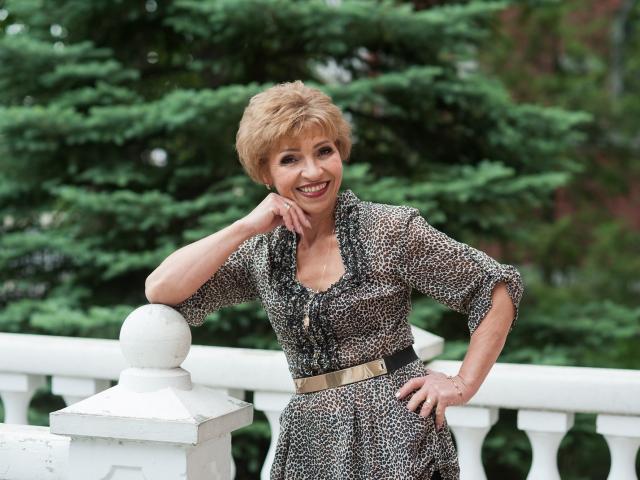 EllaBonito with short, curly blonde hair, smiling, wearing a leopard print dress, black belt, and white railing in foreground, leaning on ball, hand on hip.