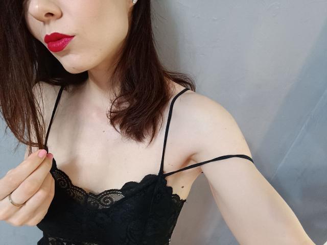AliceSunset with red lipstick, black lace bra, and pink nail polish, pulling down a thin strap on her black lace top, showing her small tits. Her face is partially visible.