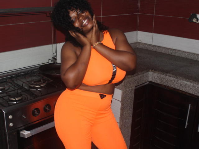 Annykaa with dark skin, curly hair, smiling, wearing orange sports bra and pants, hand on cheek, standing in kitchen, showing large boobs and curvy ass.