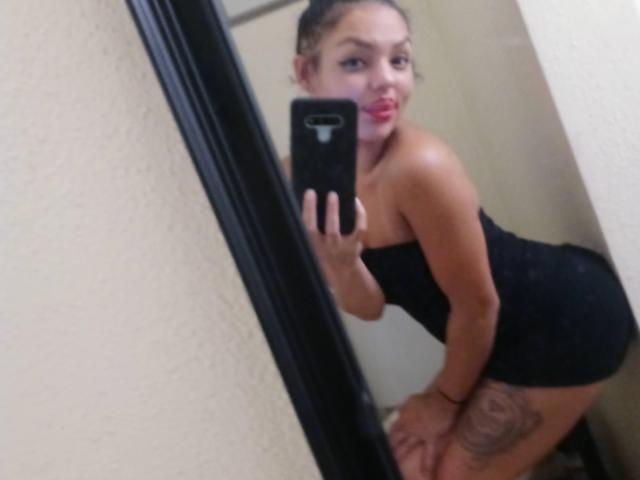 NaughtyNikkix with her ass in a tight black dress, leaning forward, taking a mirror selfie. She's got red lipstick, a tattoo on her thigh, and a phone with a white logo.