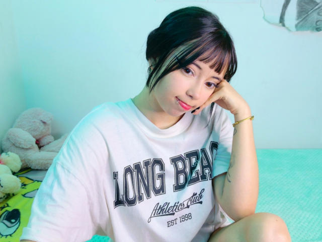 Arieyaara with short black hair and bangs, wearing a white "Long Beach" sweatshirt, sitting on a green bed, head resting on her hand, smiling gently. Soft lighting.