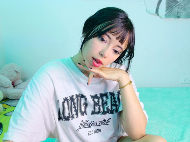 Arieyaara with short black hair, wearing a white "Long Beach" shirt, resting chin on hand, pink lipstick, soft green bed, beige stuffed toy in background, slightly parted pink lips.