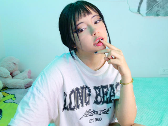 Arieyaara with black hair and bangs, wearing a white Long Beach shirt, sucking on her finger with pink lipstick, sitting on a green bed, looking at the camera.
