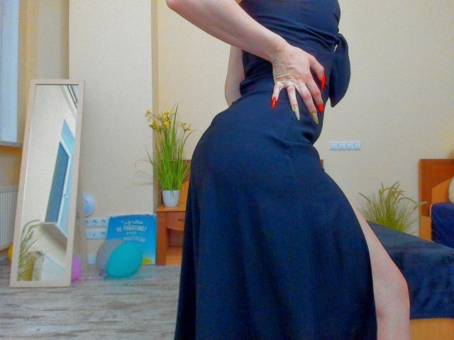 Jumiera's black dress hugs her curvy ass, showing a high thigh slit. She's wearing long, red, and white manicured nails, standing with one hand on her hip.