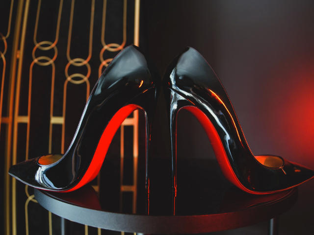Shiny black stiletto heels with red soles, standing on a round table, reflecting light, showing their glossy, curved upper parts, and pointed toes. LongetsLegs-hot's sexy shoes.