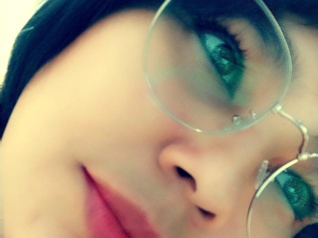 PrittyAlicia's face with green eyes, wearing round glasses, has pink lips slightly parted. Her black hair is visible on the left side of the image.