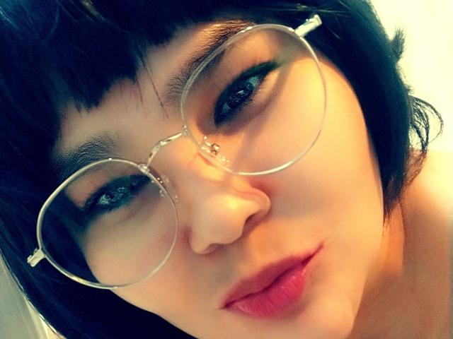 PrittyAlicia has short black hair, wearing round glasses, with dark eyeliner and red lipstick. Her pale skin and slightly pouty lips are visible. Her nose is small and cute.