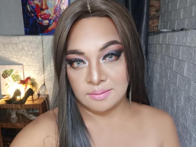VogaQueen has light brown skin, blue eyes, and long brown hair. She's wearing pink lipstick, black eyeliner, and silver earrings. Her tits are not visible. She's looking at the camera.