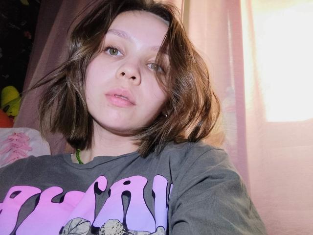 VanessaKit-hot with shoulder-length brown hair, light skin, and pink lips, wearing a gray shirt with pink lettering, looking slightly to the side in a soft light.