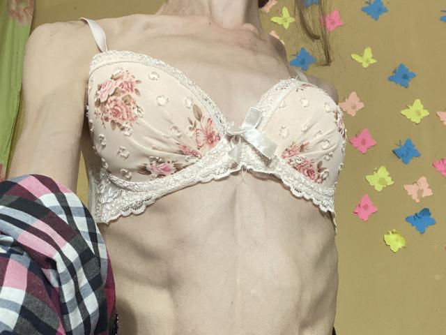 Kabblashas kleine, helle Brüste in einem floralen Spitzen-BH, die ihren Brustkorb und ihren flachen Bauch zeigen, wobei ein rosa und schwarzes Karoshirt sie teilweise bedeckt..