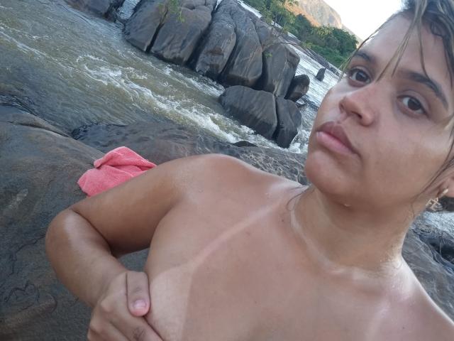 Kinha69, topless, with tan lines and wet skin, covering small tits with one hand, pink towel on rock, river flowing behind, serious expression, blonde hair, slight tan, outdoors.
