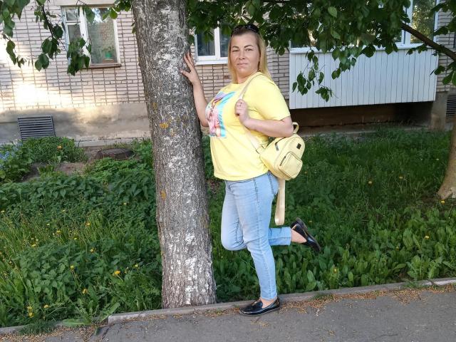NiceCaroline with medium tits, blonde, wearing yellow tee, blue jeans, black flats, yellow bag, leaning on tree, right leg raised, grassy yard, daytime, smiling.