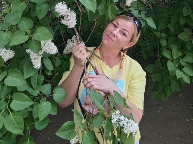 NiceCaroline with blonde hair, yellow shirt, and sunglasses on head, holding a white flower branch, smiling slightly, surrounded by green leaves and white flowers.