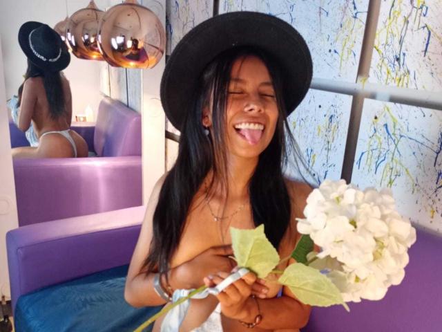 LuccyHot69 with long black hair, black hat, smiling, tongue out, holding white hydrangeas, covering small tits with hands, mirror reflection shows butt in white thong, purple sofa.