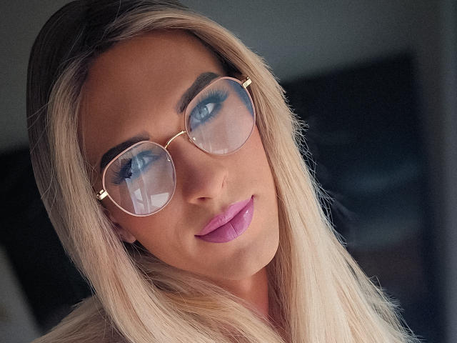 KinkyNora heeft lang blond haar, blauwe ogen en draagt ronde brillen met roze lippenstift. Haar wenkbrauwen zijn dik en ze glimlacht lichtjes. Haar borsten zijn niet zichtbaar..