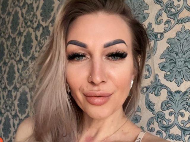 DennyBrenda has blonde hair, thick dark eyebrows, long eyelashes, and full pink lips with a subtle smile. She's wearing a nose ring and white earrings.