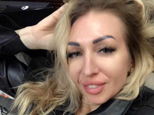 DennyBrenda with blonde hair, thick eyebrows, and full lips, lying in a car, wearing a black leather jacket, smiling slightly, hand in hair.