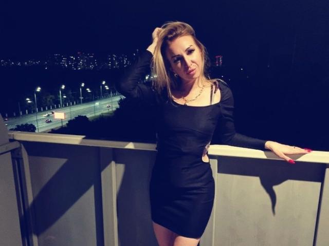 DennyBrenda está em uma varanda à noite, vestindo um vestido preto justo, com a mão no cabelo, exibindo seus seios e um sorriso adorável..