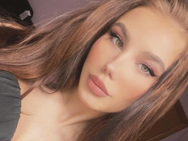 NicoleNight has long brown hair, green eyes, and full pink lips. She's wearing black, with a small mole below her right eye. Her makeup is subtle, with dark eyeliner.